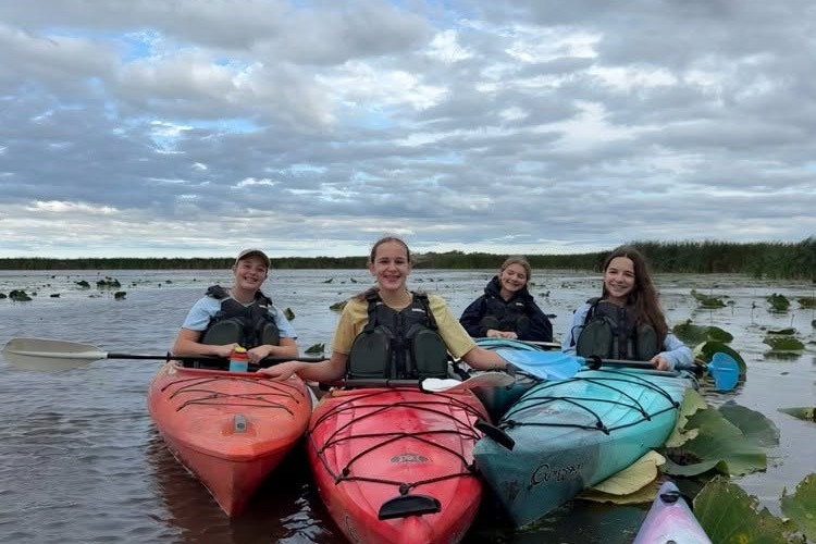 Four people in life jackets and kayaks on calm water. One kayak is red, one is blue, and two are green.