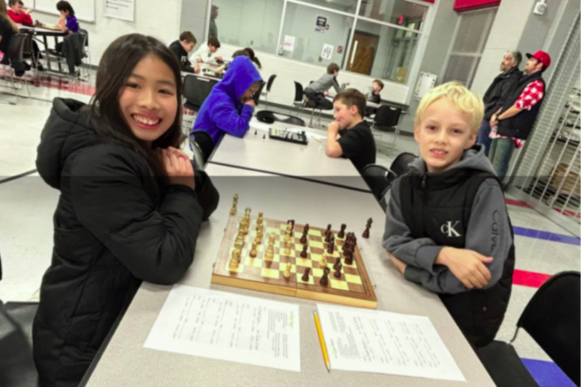 Two children sit opposite each other at a table, smiling and facing a chess set.