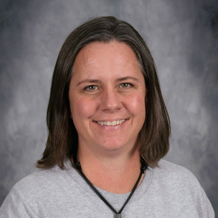 A woman with short dark hair wearing a gray shirt with a rocket and text stands against a gray background.