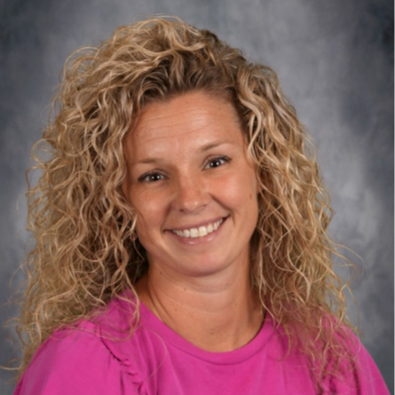 Woman with curly blonde hair wearing a pink shirt. She is smiling and looking forward. Background is gray.