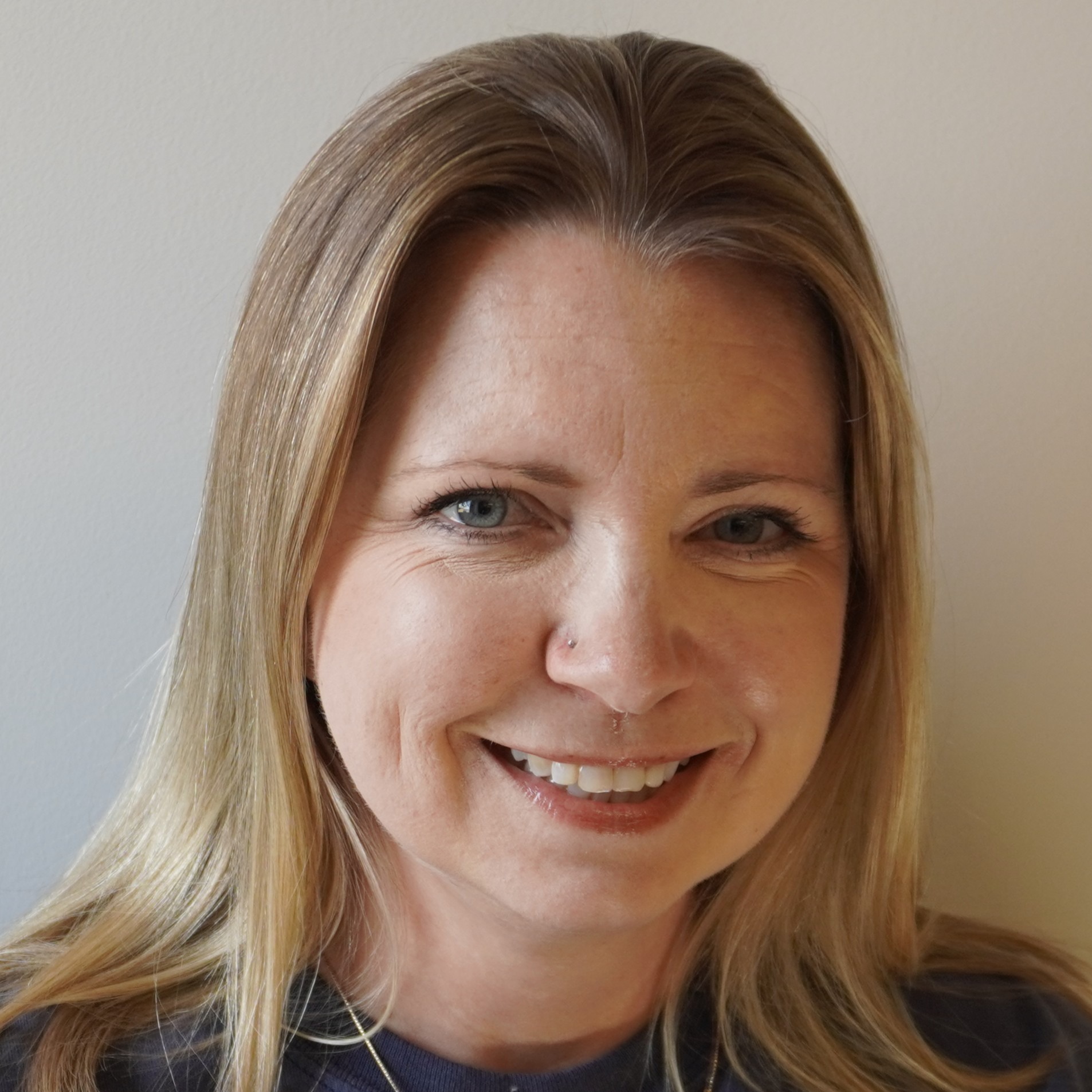 Woman with shoulder-length blonde hair wearing a navy blue shirt and a gold necklace. She is smiling.