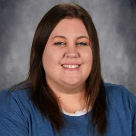 A woman with long, dark hair, wearing a blue shirt, smiles at the camera. She stands against a plain gray background.