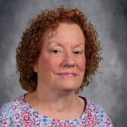 Woman with curly red hair wearing a blue and pink floral shirt against a gray background.