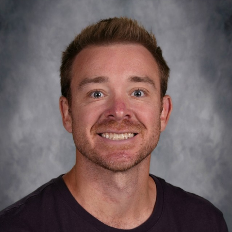 A man with brown hair, short beard, and a smile stands against a gray background, wearing a black t-shirt.