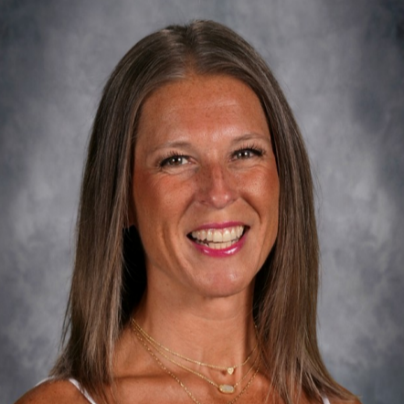 A smiling woman wearing a white tank top and multiple necklaces stands against a gray background.
