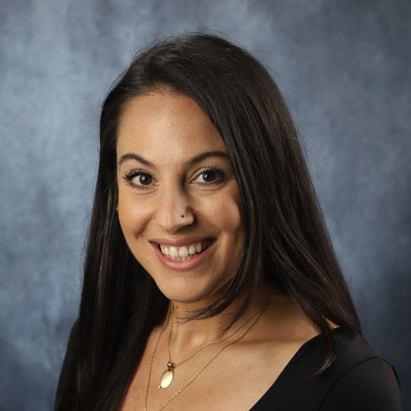 A woman with long dark hair wearing a black top and multiple necklaces, smiling and posing for a photo.