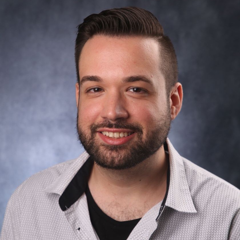 A man in a white patterned shirt with a black collar and a black shirt underneath. He has a beard and is smiling.