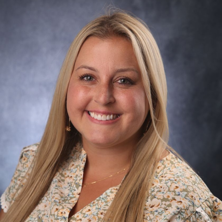 Blonde-haired woman with a floral shirt, smiling at the camera against a light gray background.