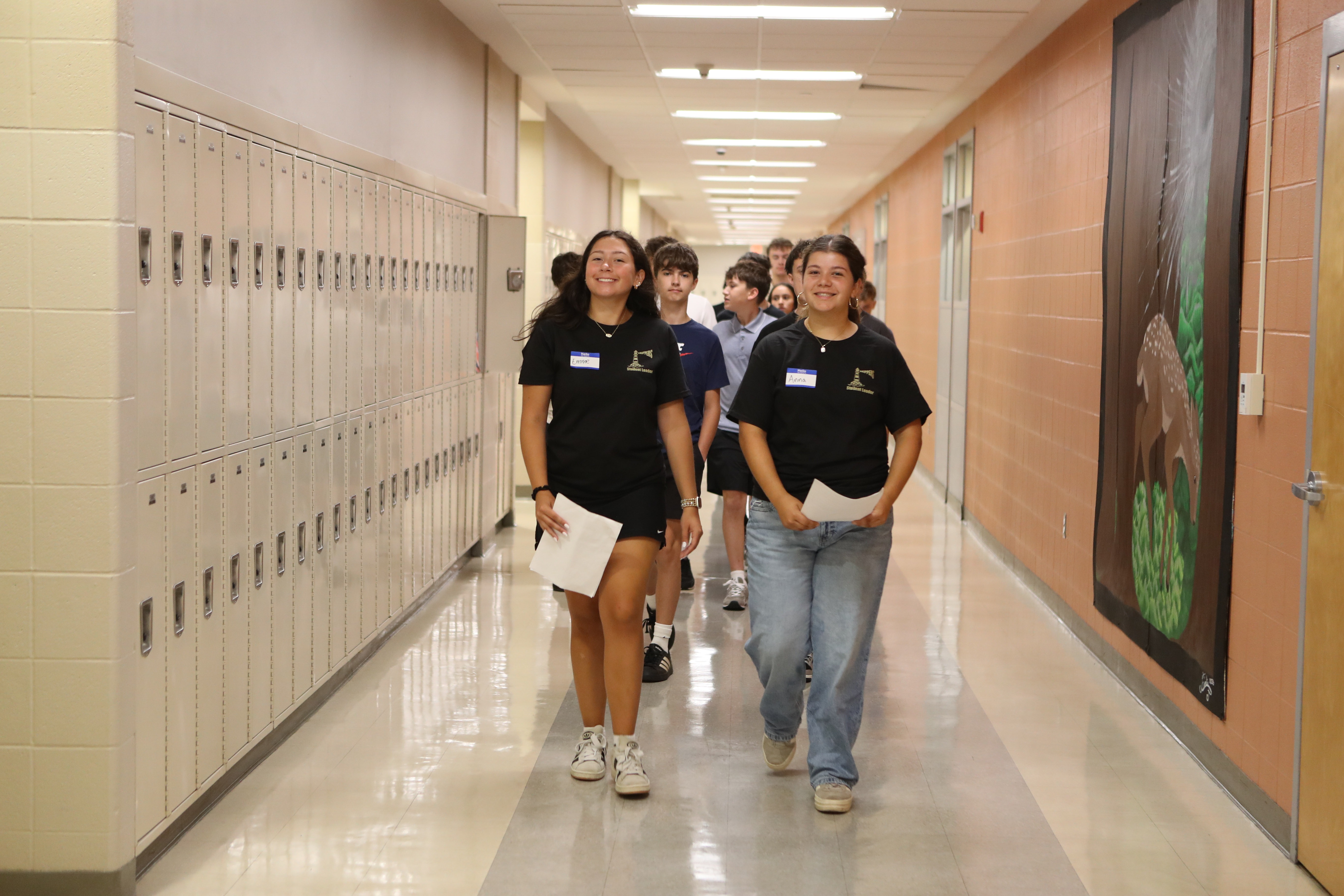 A hallway with lockers on the left and art on the right. Two people are walking, one holding papers.