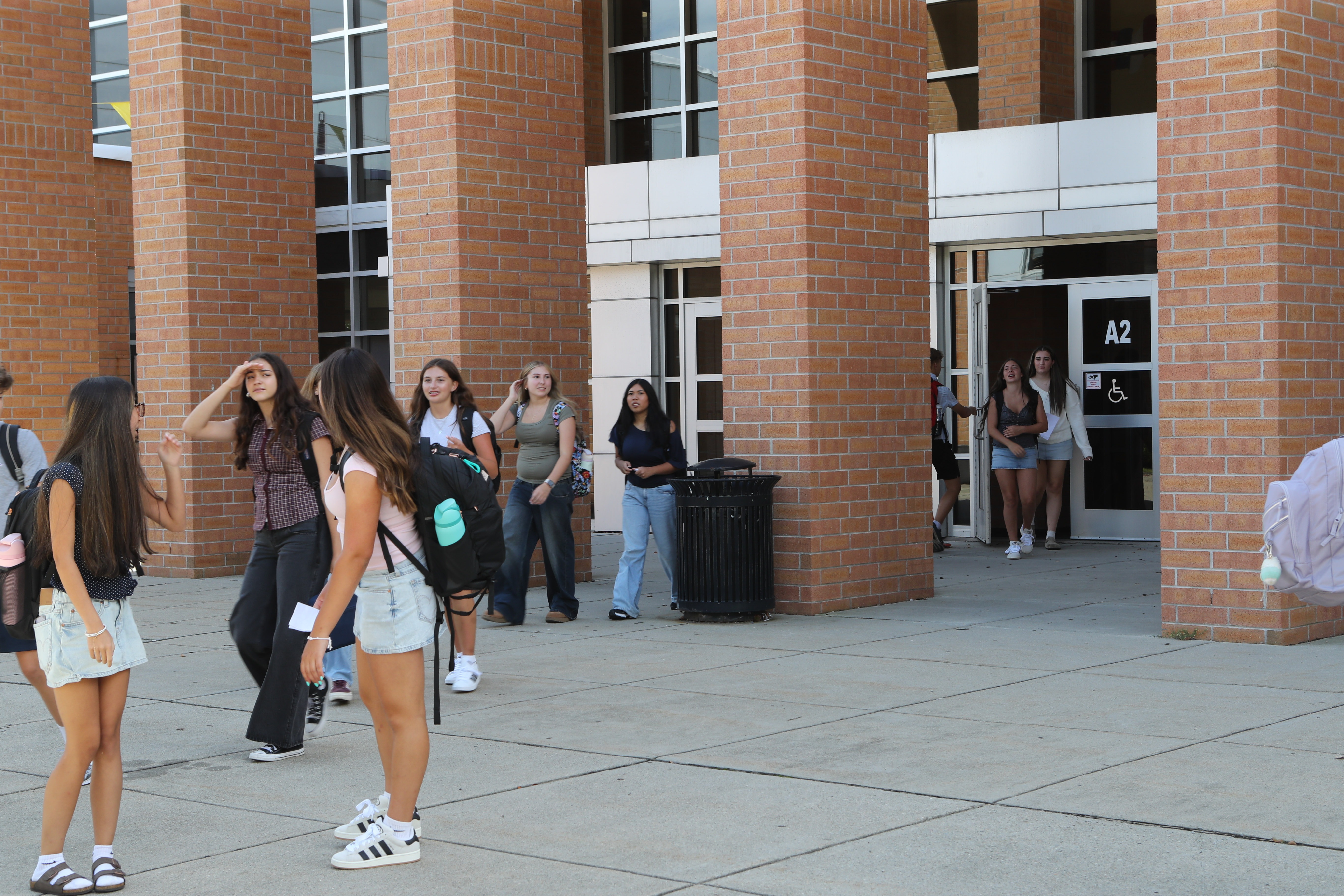People walk outside a building with brick pillars. Some carry backpacks. One wears a hat. Trash can nearby.