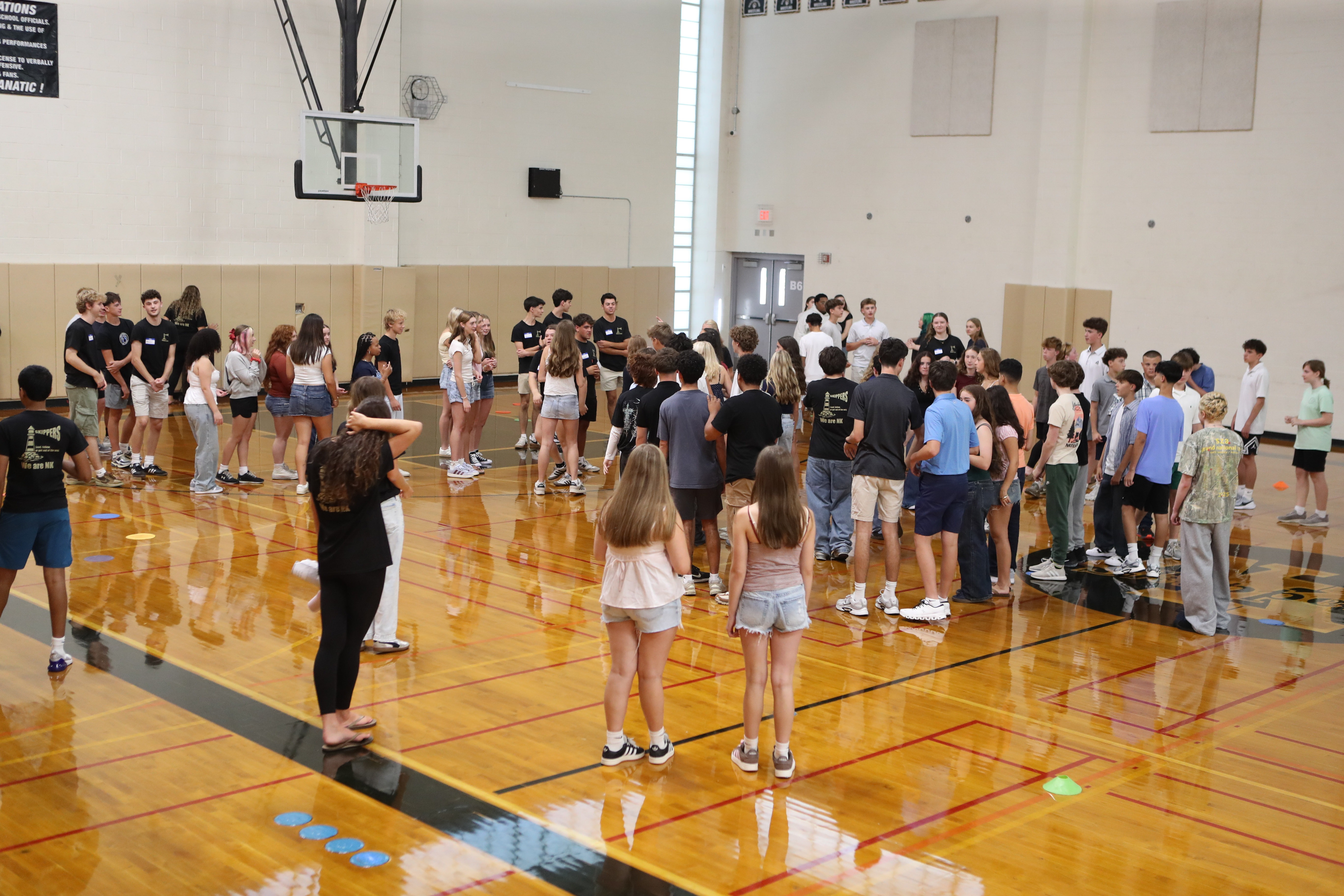A group of people gather in a gym. Some stand, others walk. The floor has blue circles.