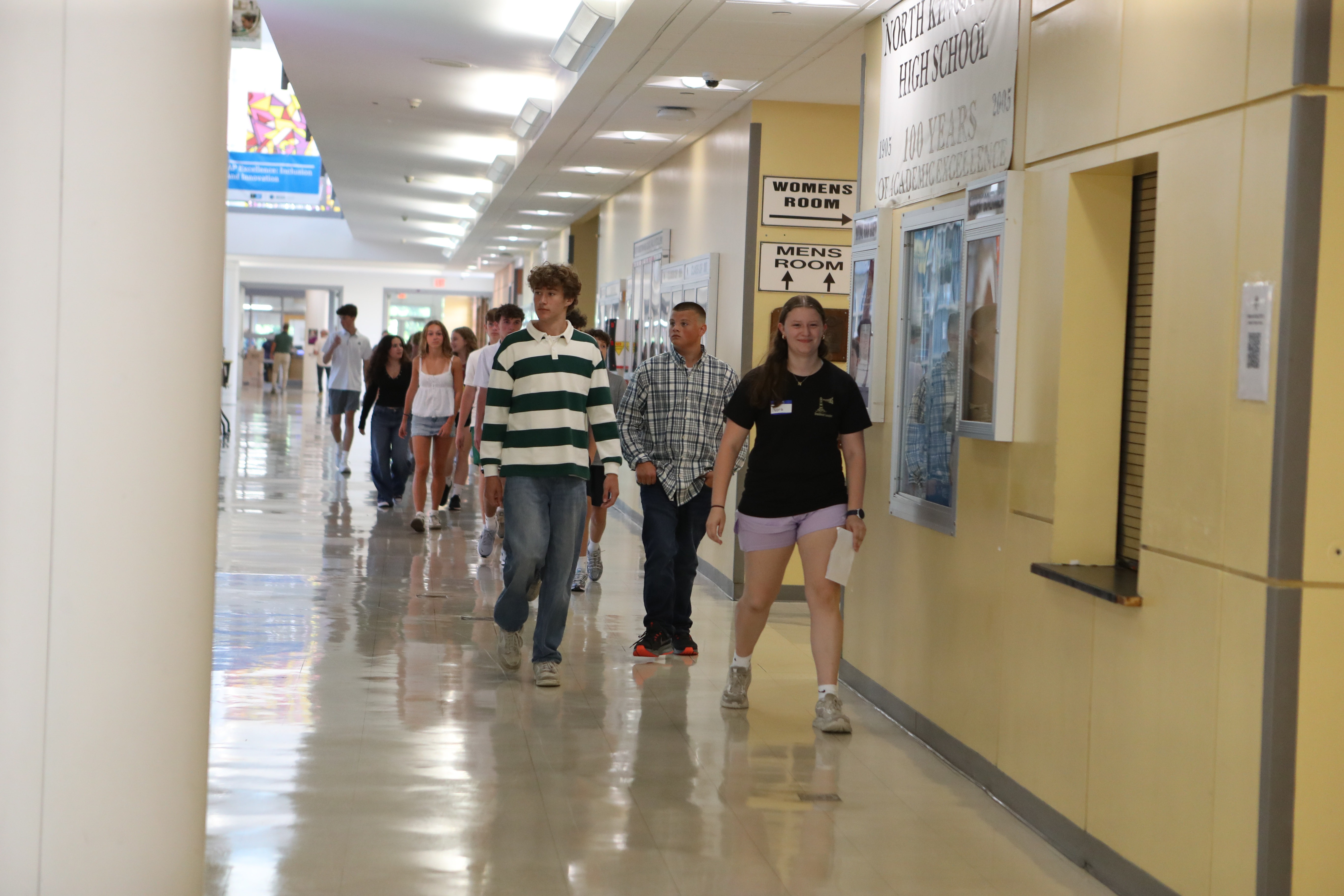 People walk in a school hallway with yellow walls and tiled floor, accompanied by others. Posters and a bulletin board adorn the walls.