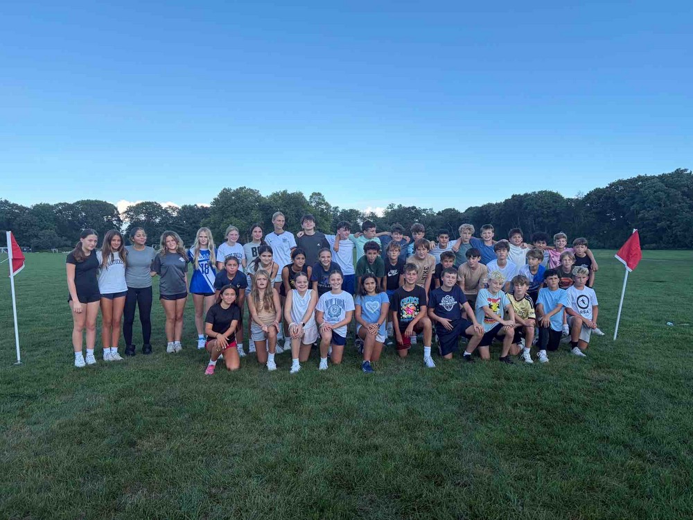 Group of people in sports gear standing and sitting on a field with two flags. Trees are visible in the background.