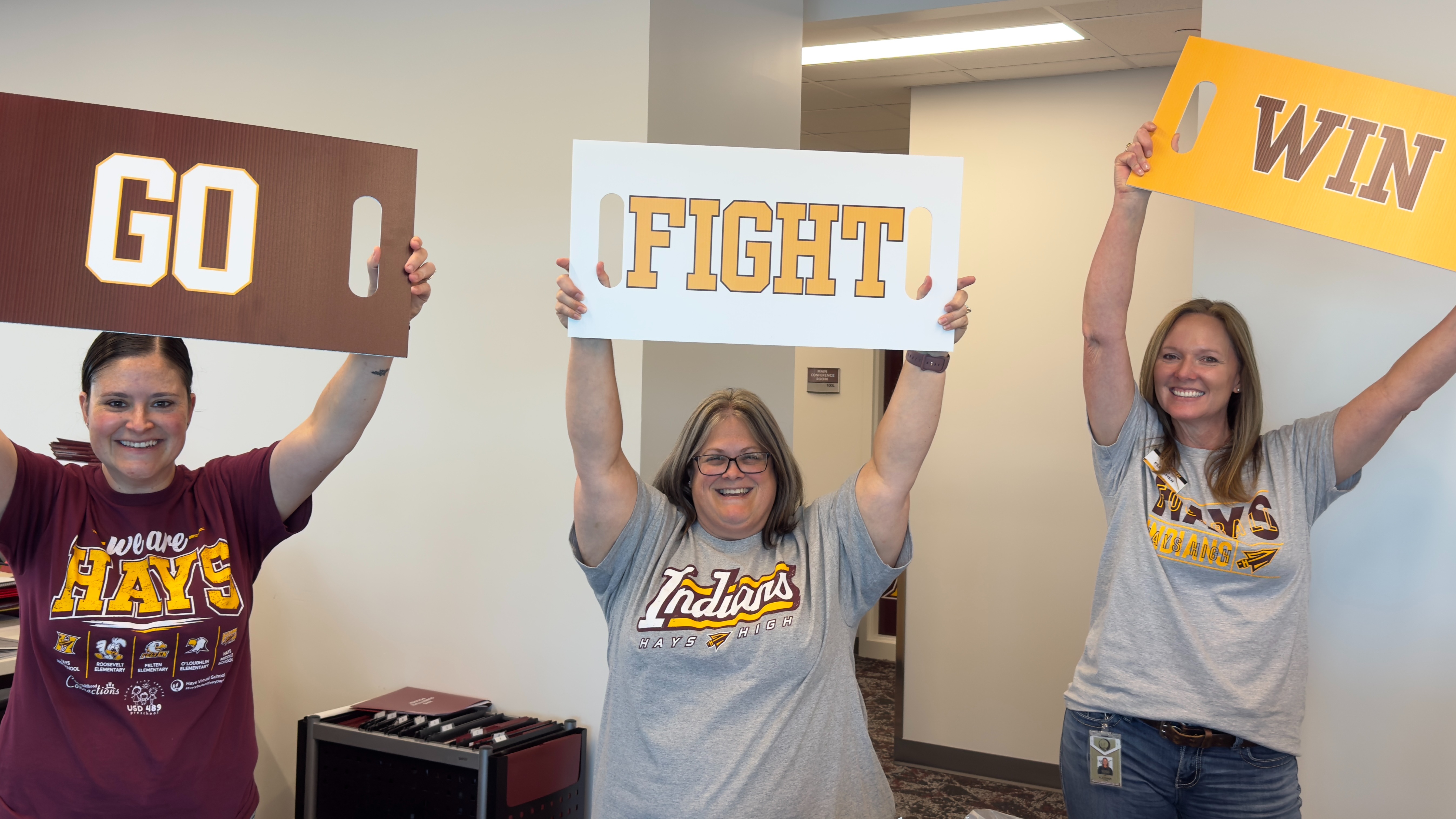 Three women hold up signs that read "I GO", "FIGHT", and "WIN" in a room.