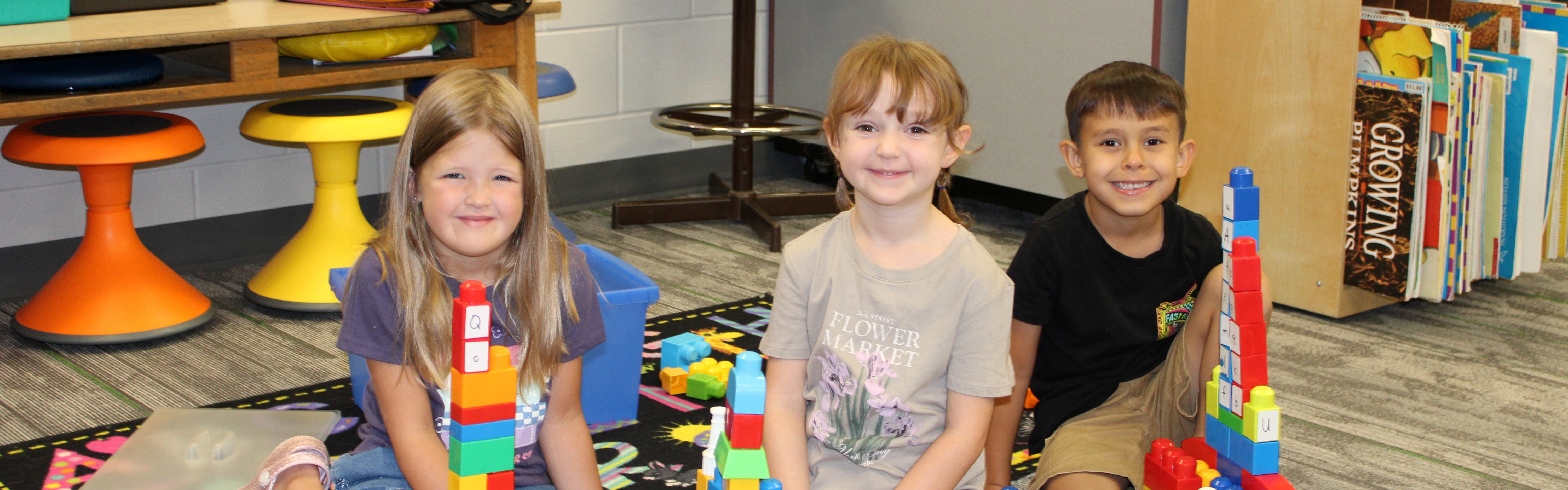 Three children sit on the floor in a classroom, playing with colorful building blocks. Behind them, a classroom with tables, chairs, and storage units is visible.