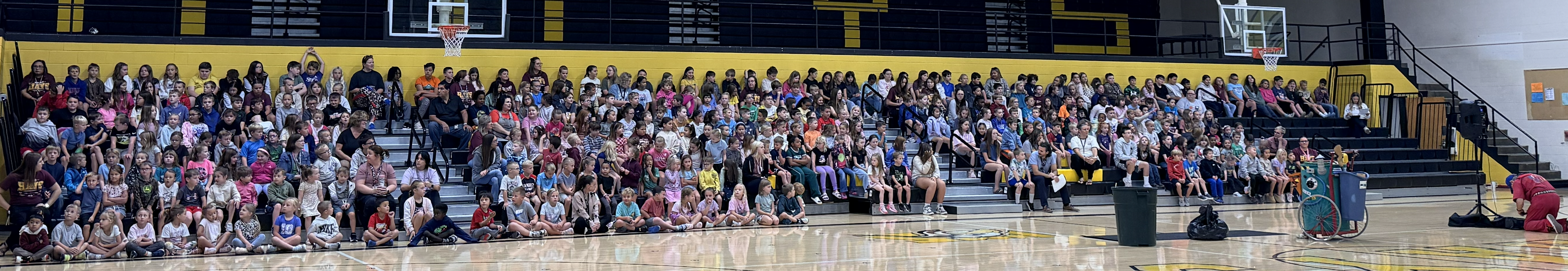 Large crowd gathered in a gymnasium, some seated and others standing, facing a stage with a person.