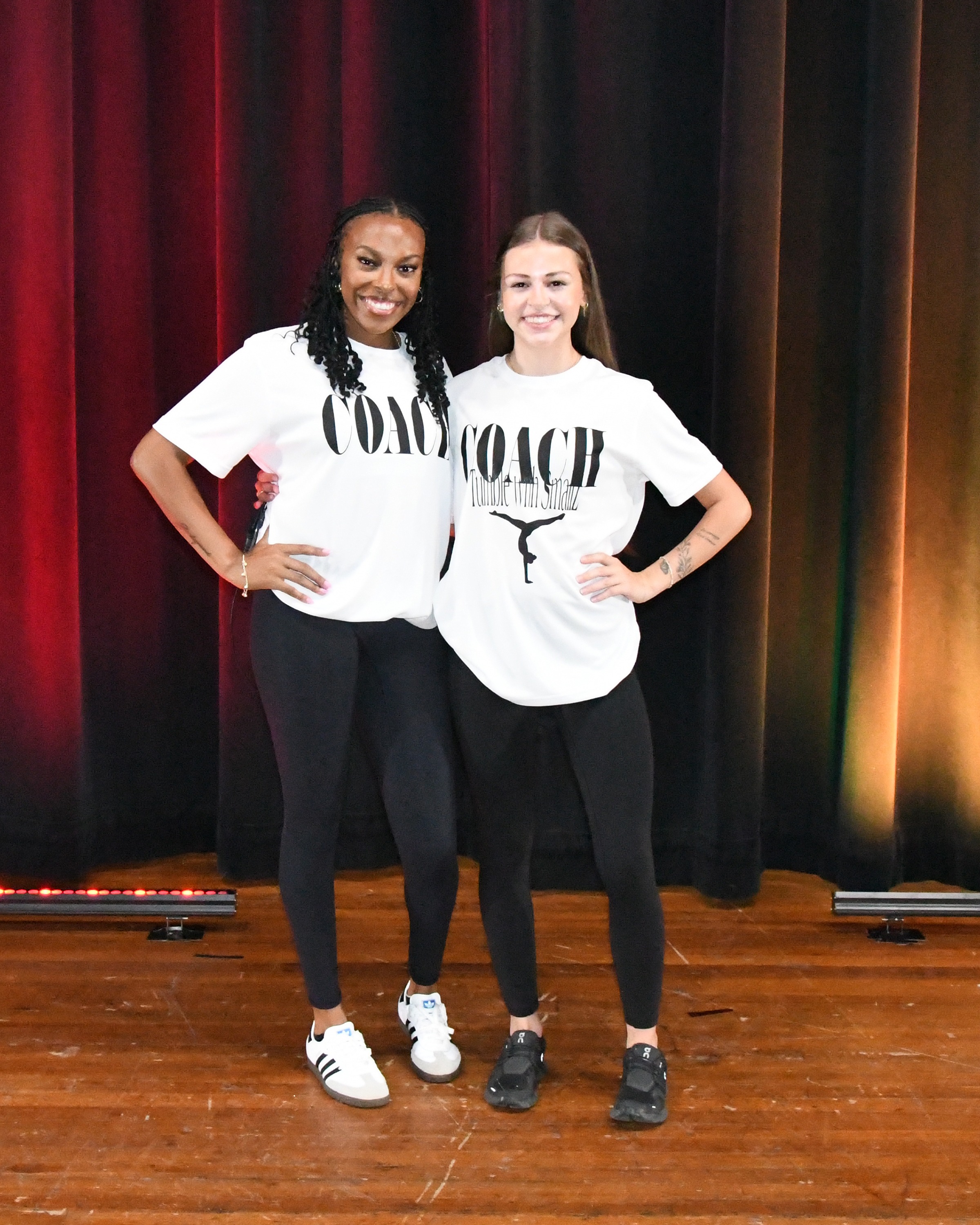 Two women in matching white "COACH" T-shirts, black leggings, and sneakers pose on a stage with a red curtain.