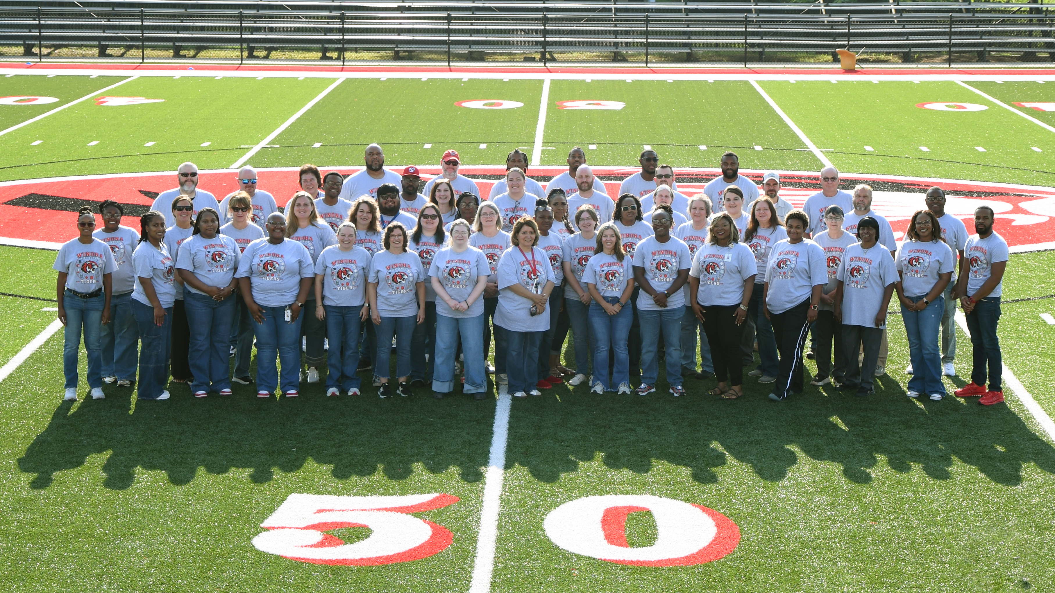 A group of people in white shirts and jeans posing on a football field with "50" marking.