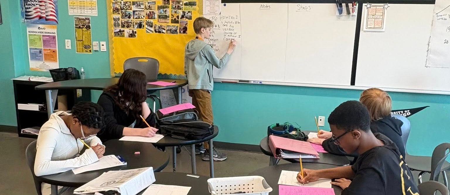 A student writes on the whiteboard while other students take notes.