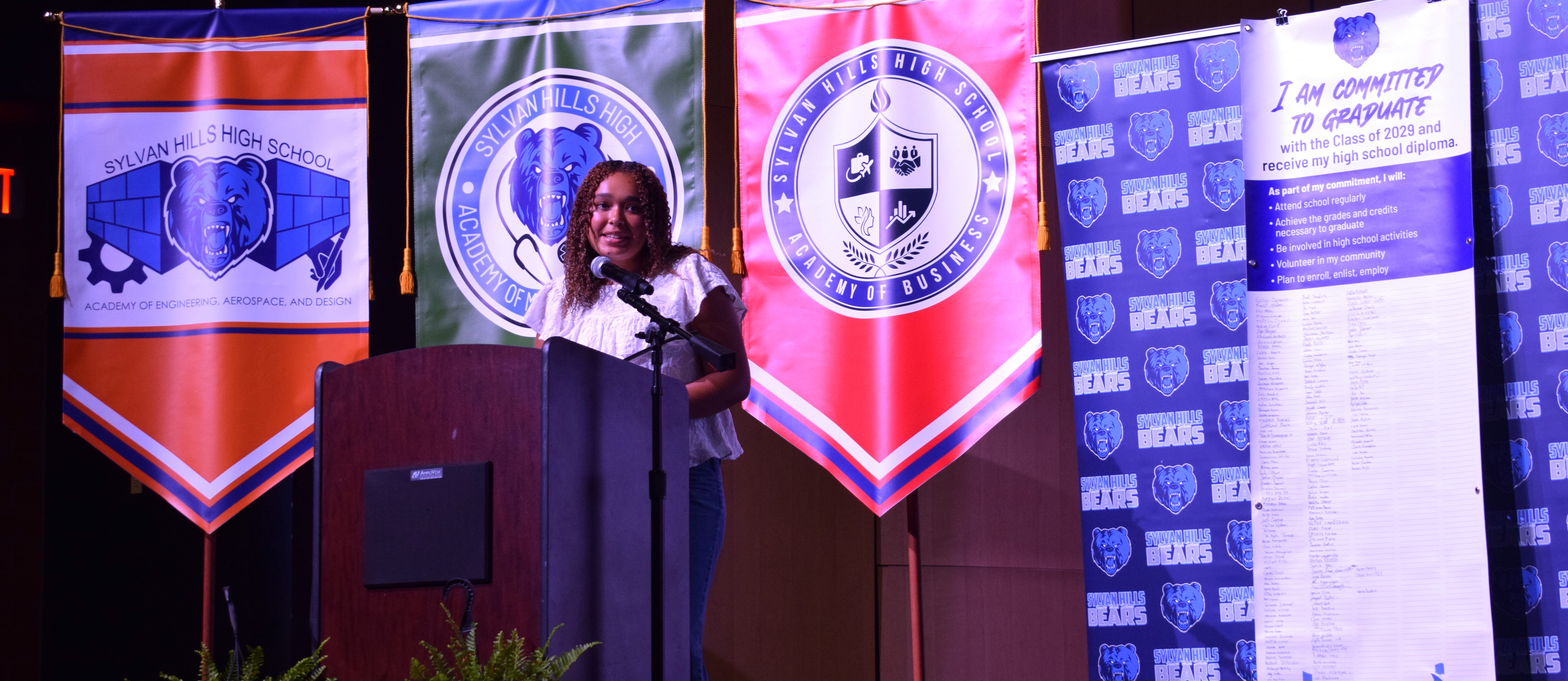 A student stands in front of Academy banners and behind a podium. A pledge to graduate banner is beside her.