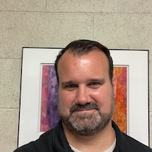 A head shot of Director Ryan Fairchild stands in front of a beige wall, a piece of art behind his head