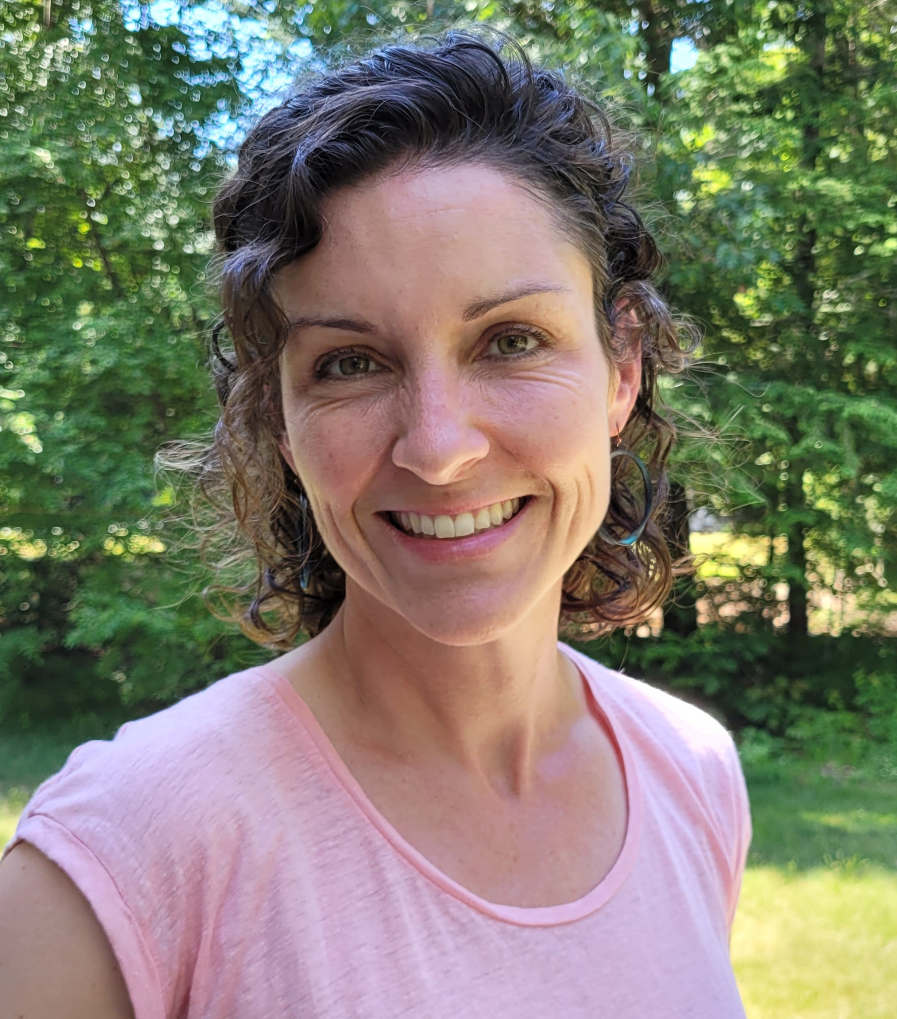 Sarah Plummer in a pink shirt stands in front of greenery outside