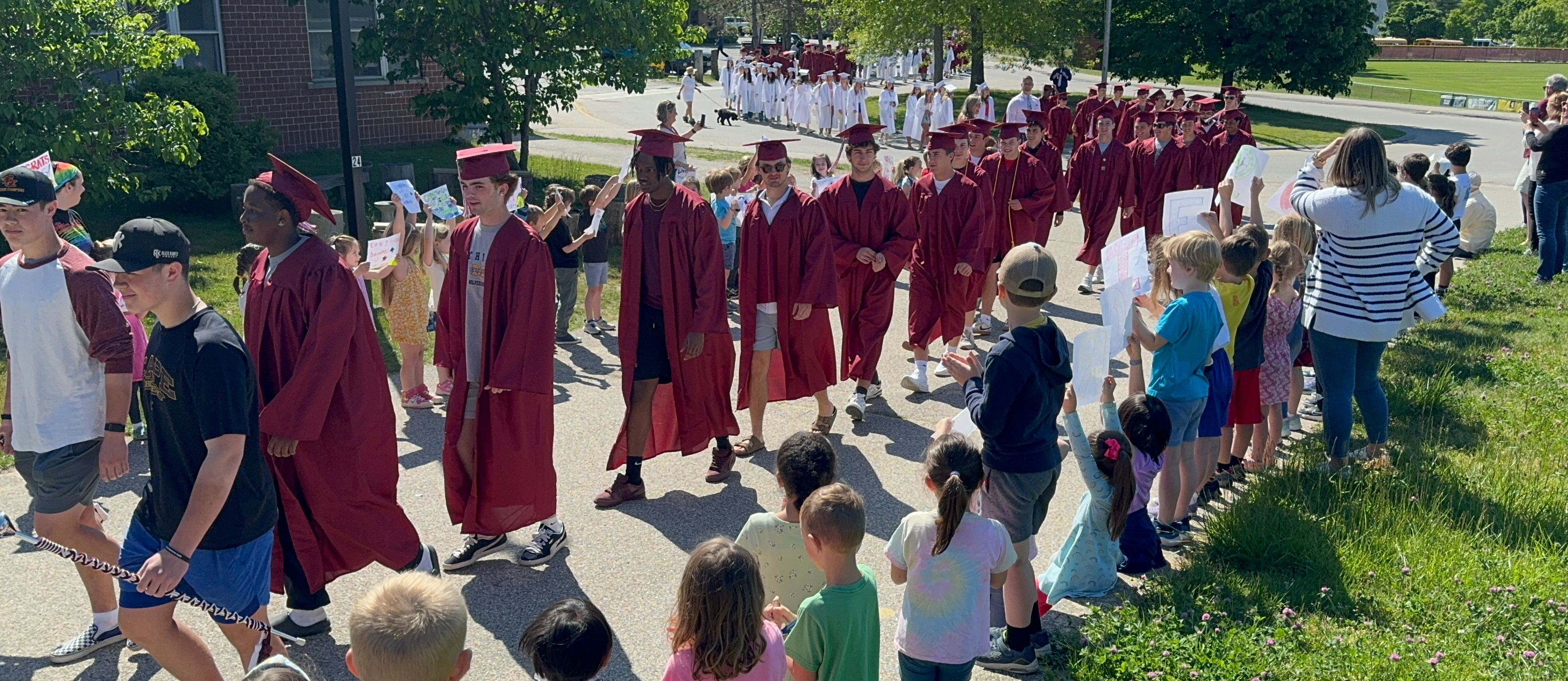 Students on the Graduation Walk of 2024. Several students walk in a line wearing caps and gowns. Along the sidewalk, on both sides, are elementary students and staff.