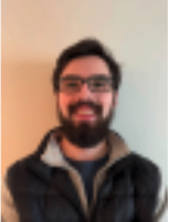 Andrew smiles in front of a plain white wall. He is wearing rectangular framed glasses . He has medium length black hair with a full beard. He is wearing a vest on top of a t-shirt. 