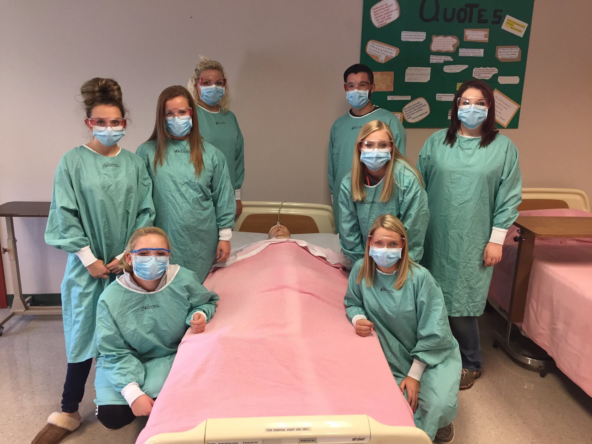 A group of CNA students in PPE stand pose near mannequin "patient" bed