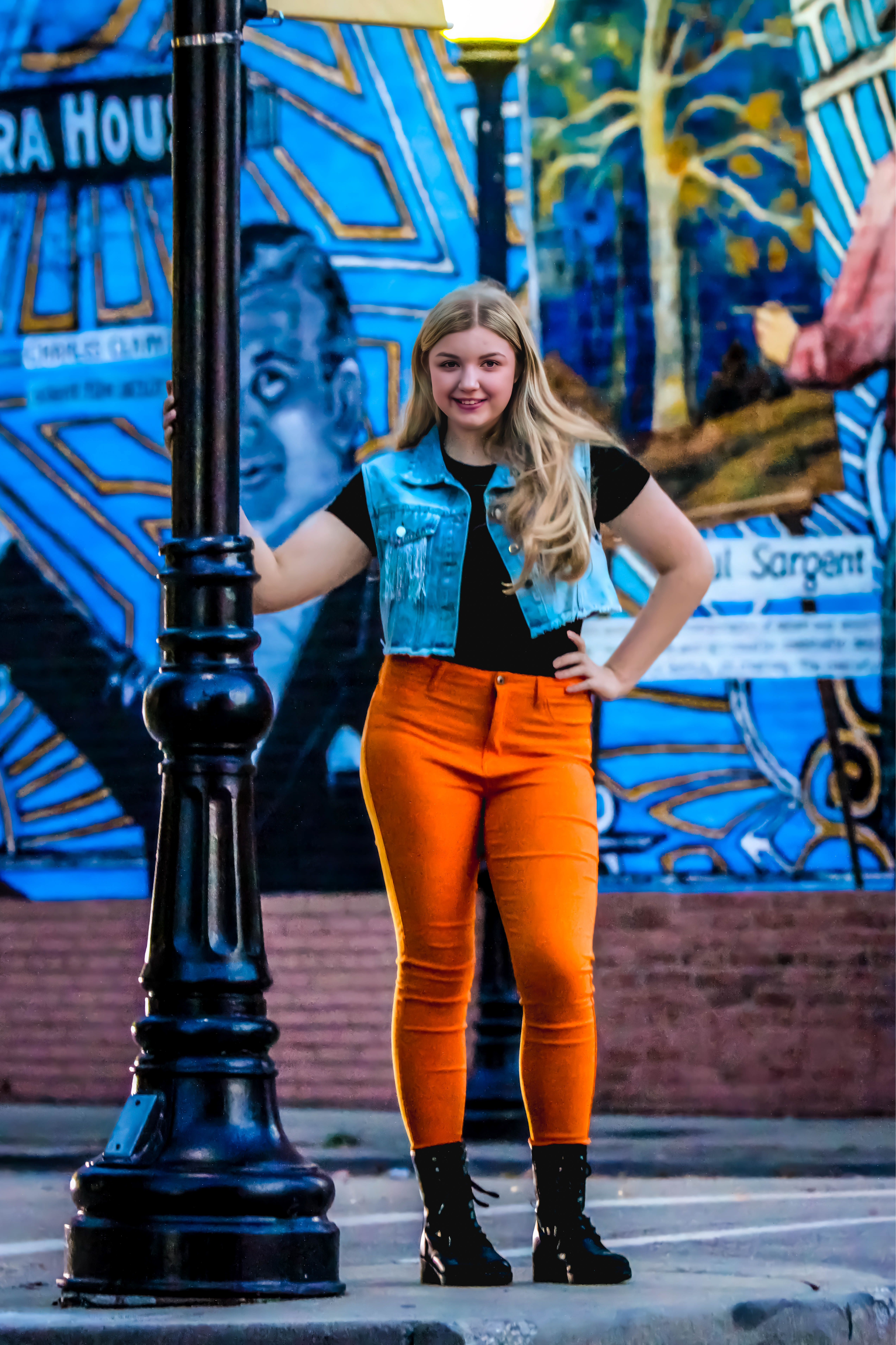 Blonde girl in bright orange pants, blue jean jacket, leaning against black pole with bright background. Faith, Freshman, Soprano.