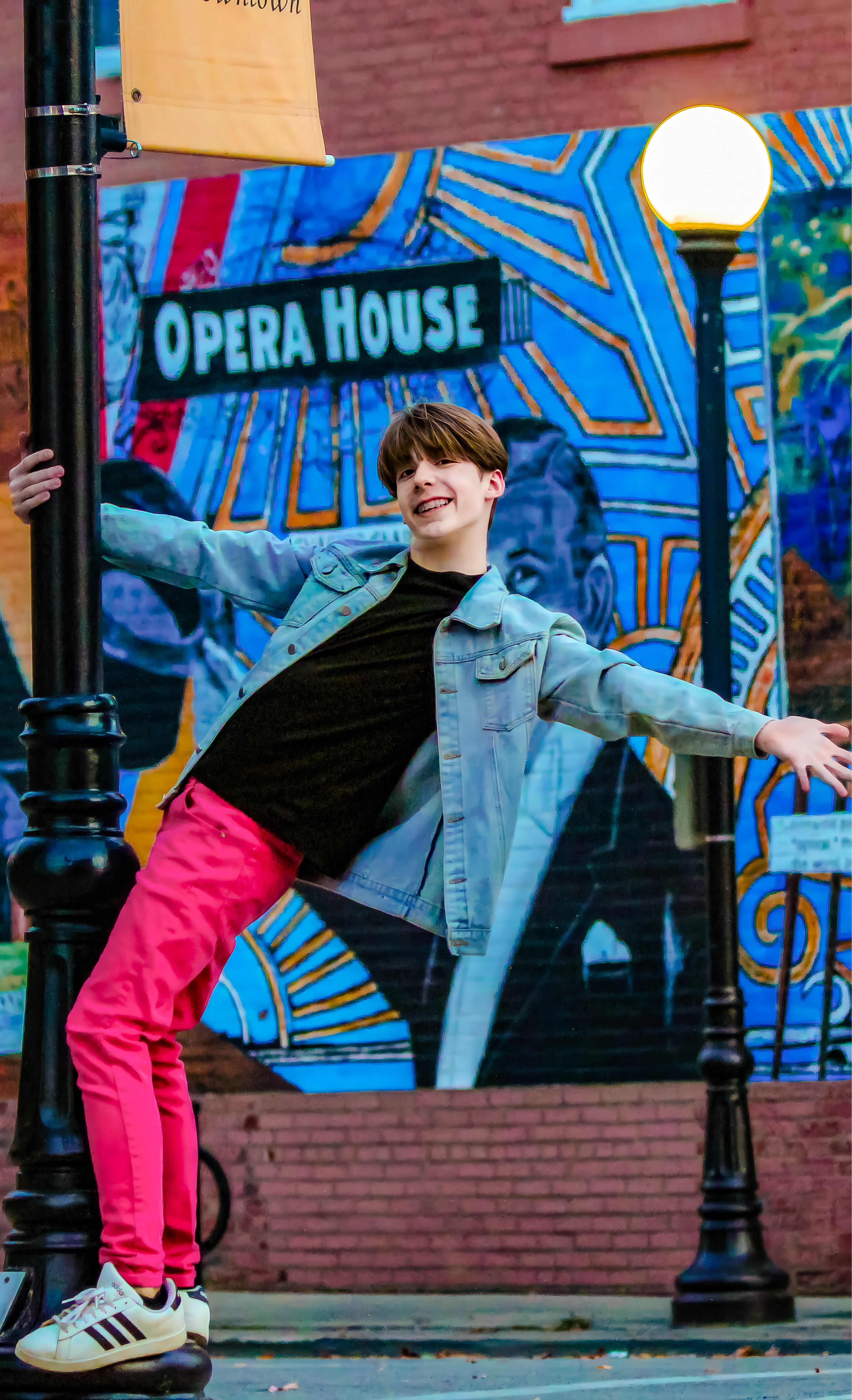 Brunette boy in bright pink pants, black shirt, and jean jacket hanging off black pole with bright background. Eli, Freshman, Tenor.