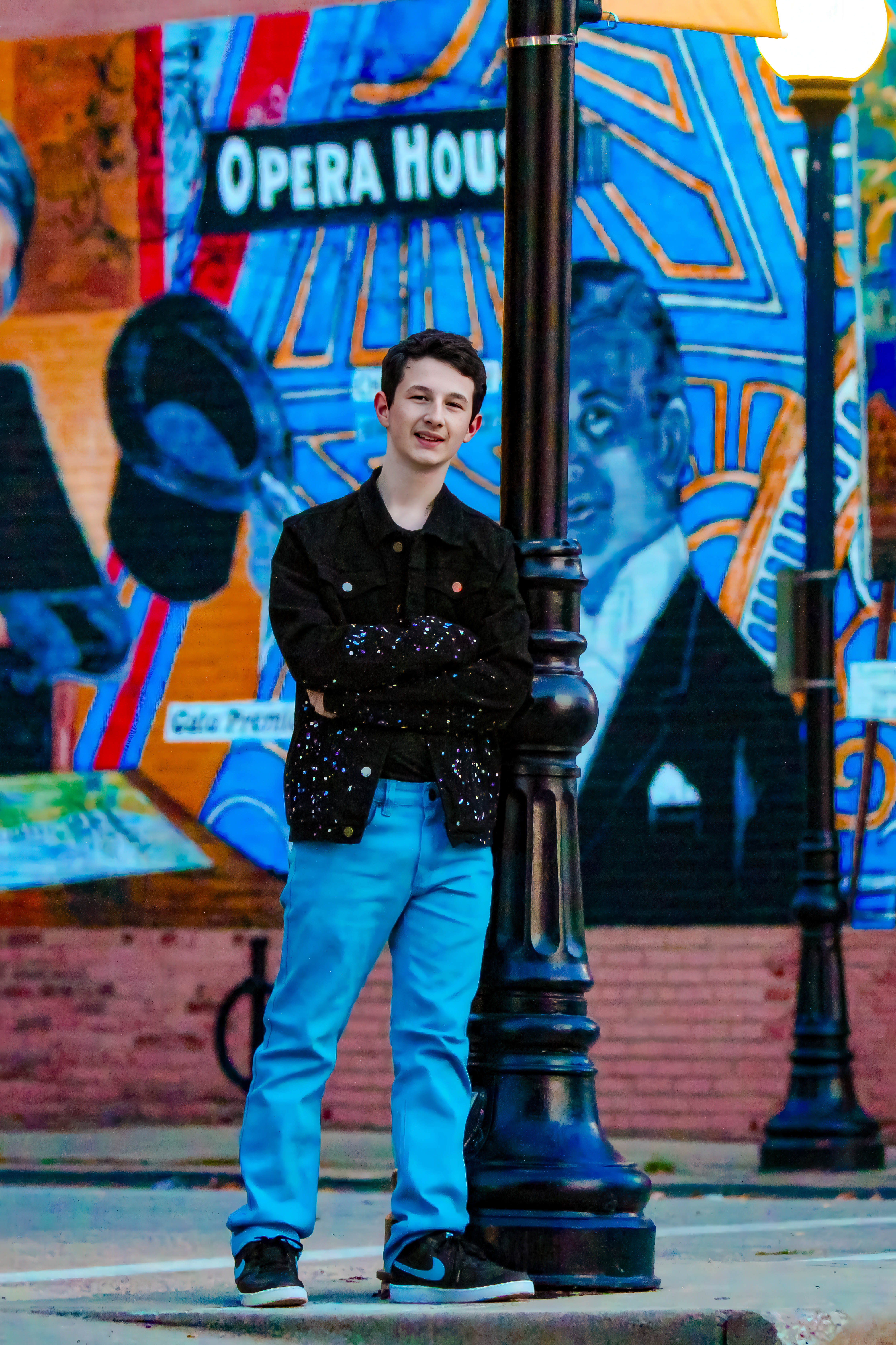 black hair boy in bright blue pants and black jacket leaning against pole with bright background. Brayden, Freshman, Bass.