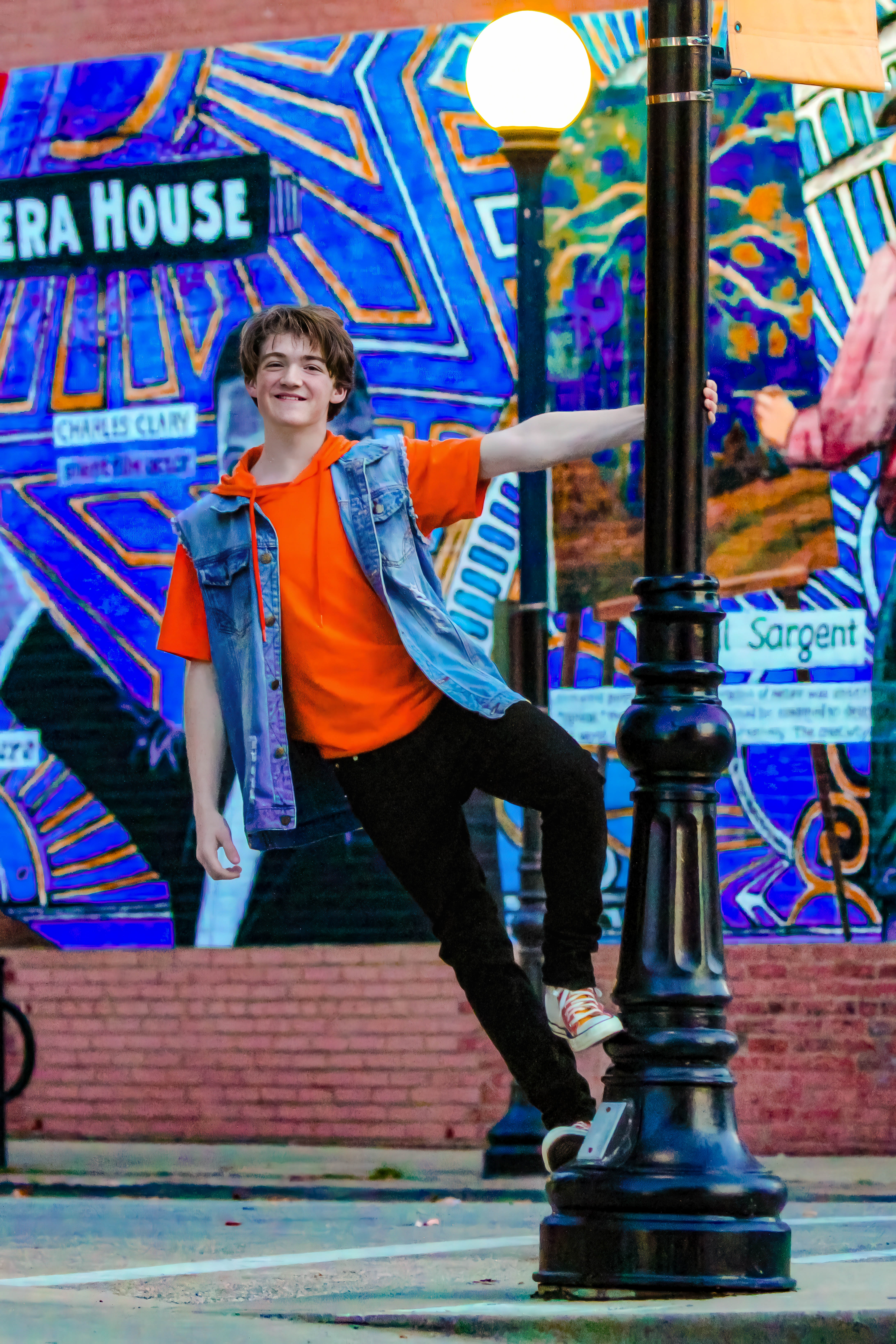 Brunette boy in bright orange top, jean jacket, and black pants hanging off black pole. Noah Northrop, Freshman, Vocal Percussionist