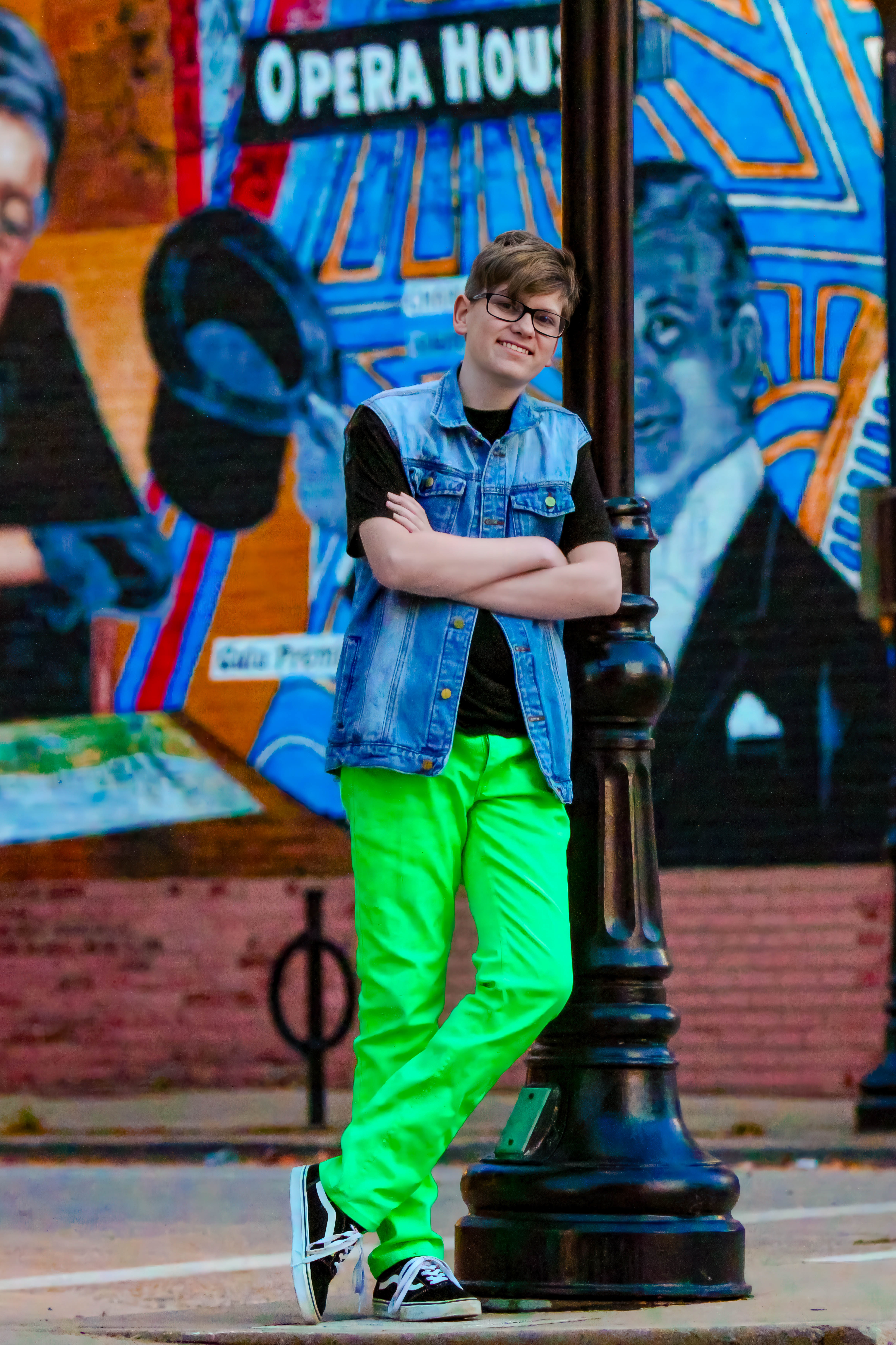 Brunette boy in jean jacket, black top, and bight green pants leaning against black pole. Sam Bates, Freshman, Tenor