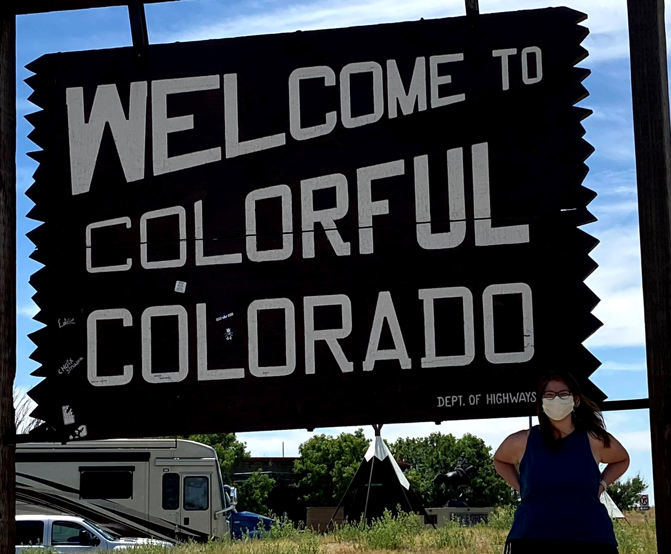 Image of  a person wearing a mask standing in front of  a sign that reads Welcome to Colorful Coloradoo