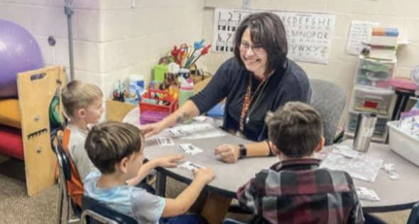 This is an image of a teacher talking to students behind a desk and working with them. 