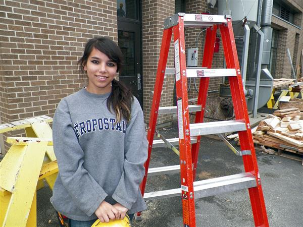 Student holds construction hat while standing next to a ladder outside
