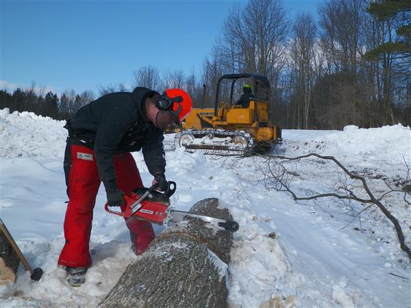 One student uses a chainsaw to cut a log and the other uses machinery to push snow