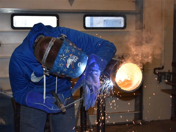 Student wlds a pipe with welding mask on as sparks fly