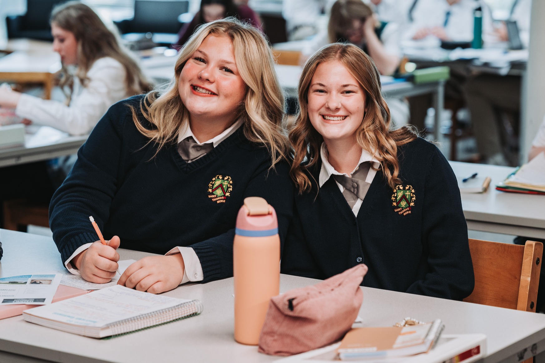 Two women in school uniforms sit at a table, smiling. One holds a pen and the other has a flask.