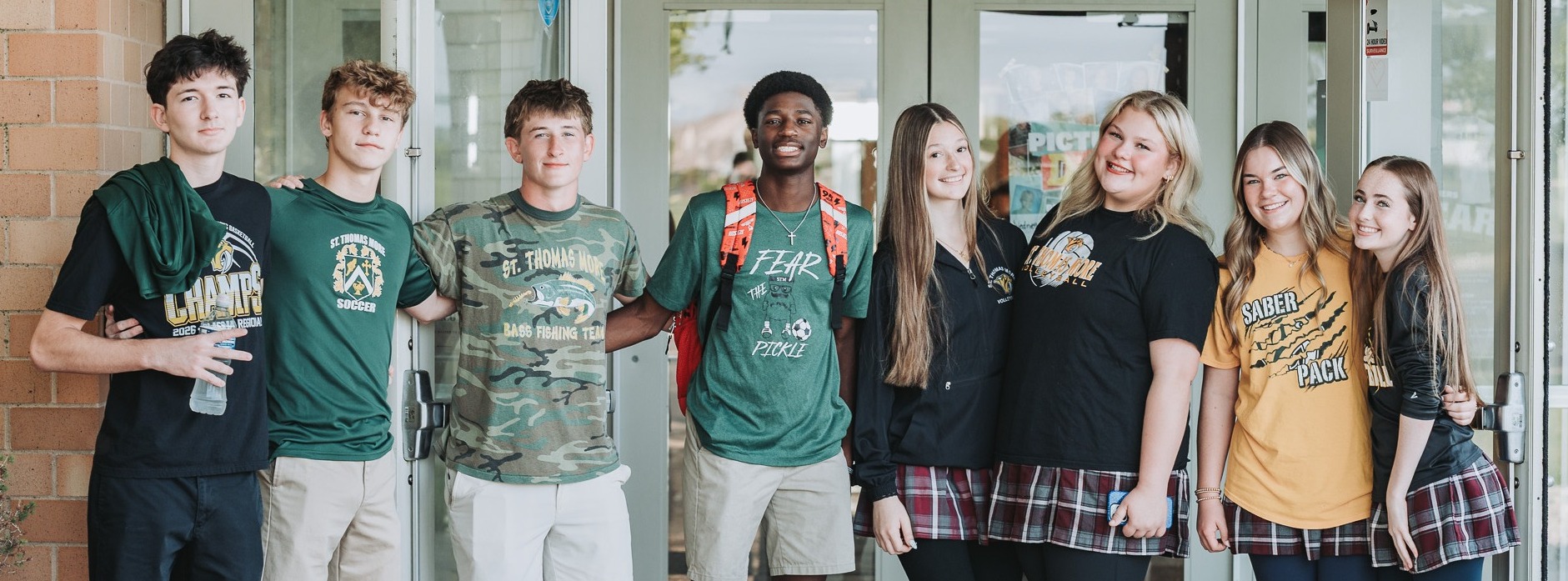 A group of seven students in various school colors pose outside a building with glass doors.