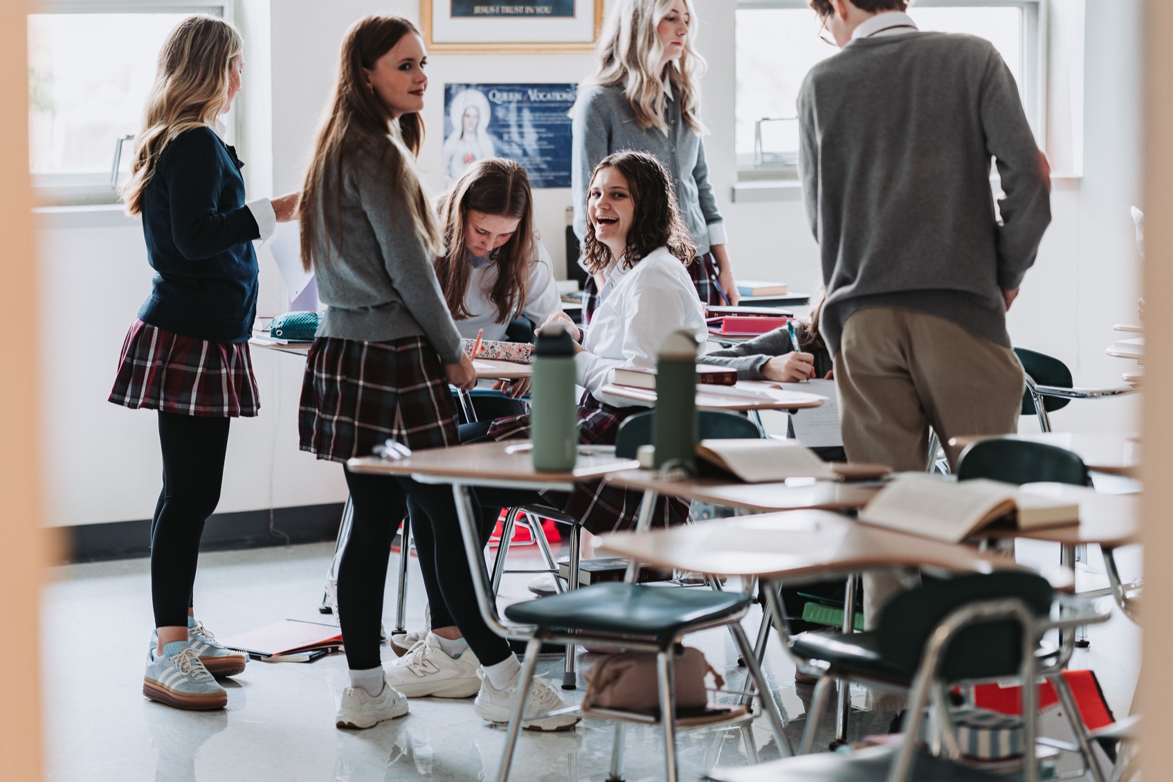 Several students gathered around desks in a classroom, one standing at the front, others seated, one leaning on a desk.