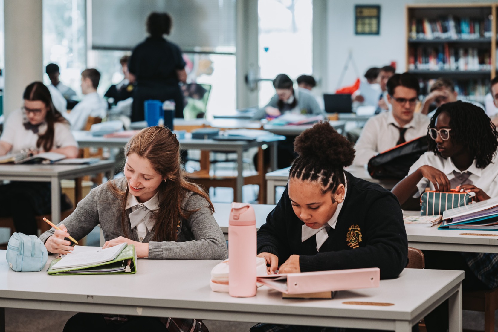 Students in a classroom setting, seated at desks, engaged in writing. Rows of desks, books, and library shelves in the background.