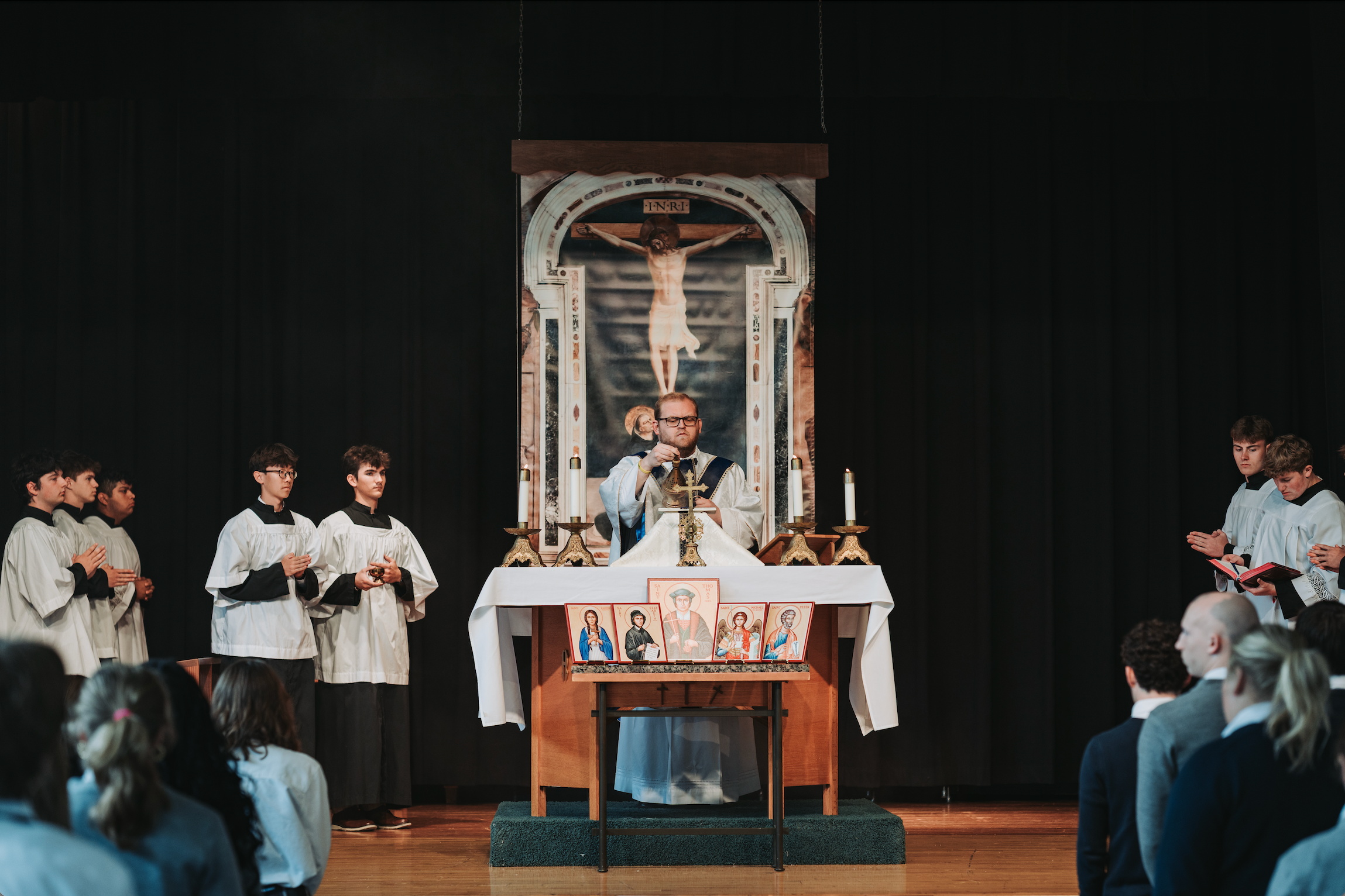 A priest leads a religious ceremony in front of a cross altar. Several individuals in white robes stand behind him.