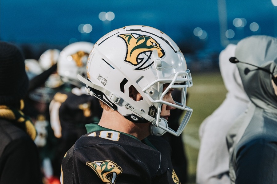Football player in white helmet with green and yellow designs looks forward on field. Teammates and others stand behind.