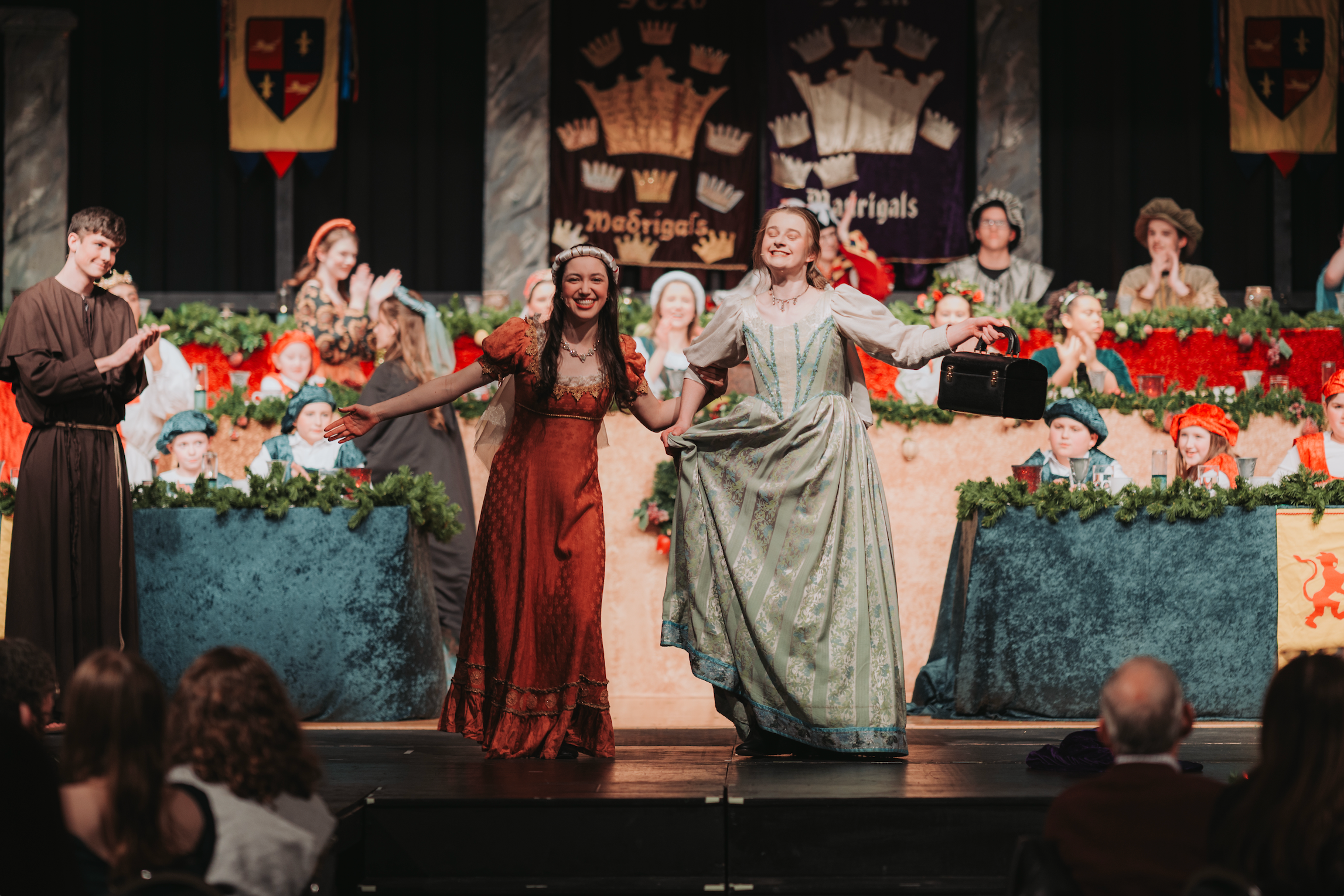 Two women in historical costumes stand on stage, one holding a purse, with others behind them and audience members watching.