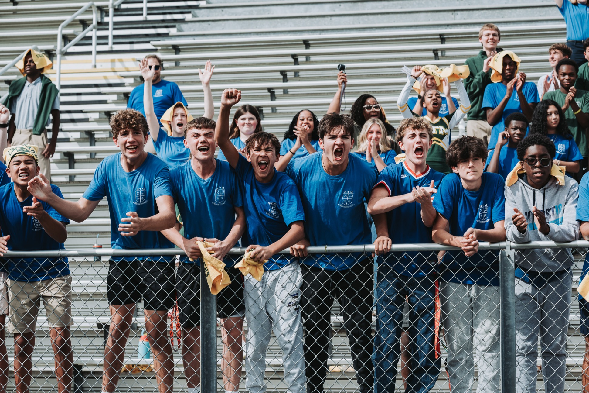 A group of young people in blue shirts and shorts cheer in an outdoor stadium. Bleachers in the background.