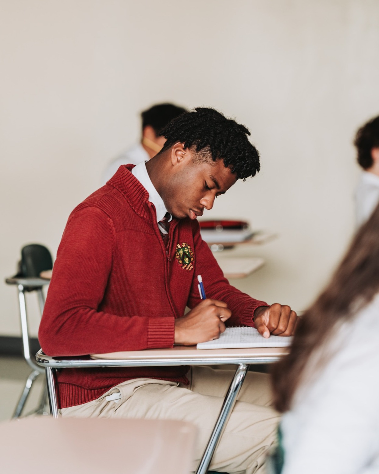 A student wearing a red sweater and tie, seated, writing in a notebook with a pen.