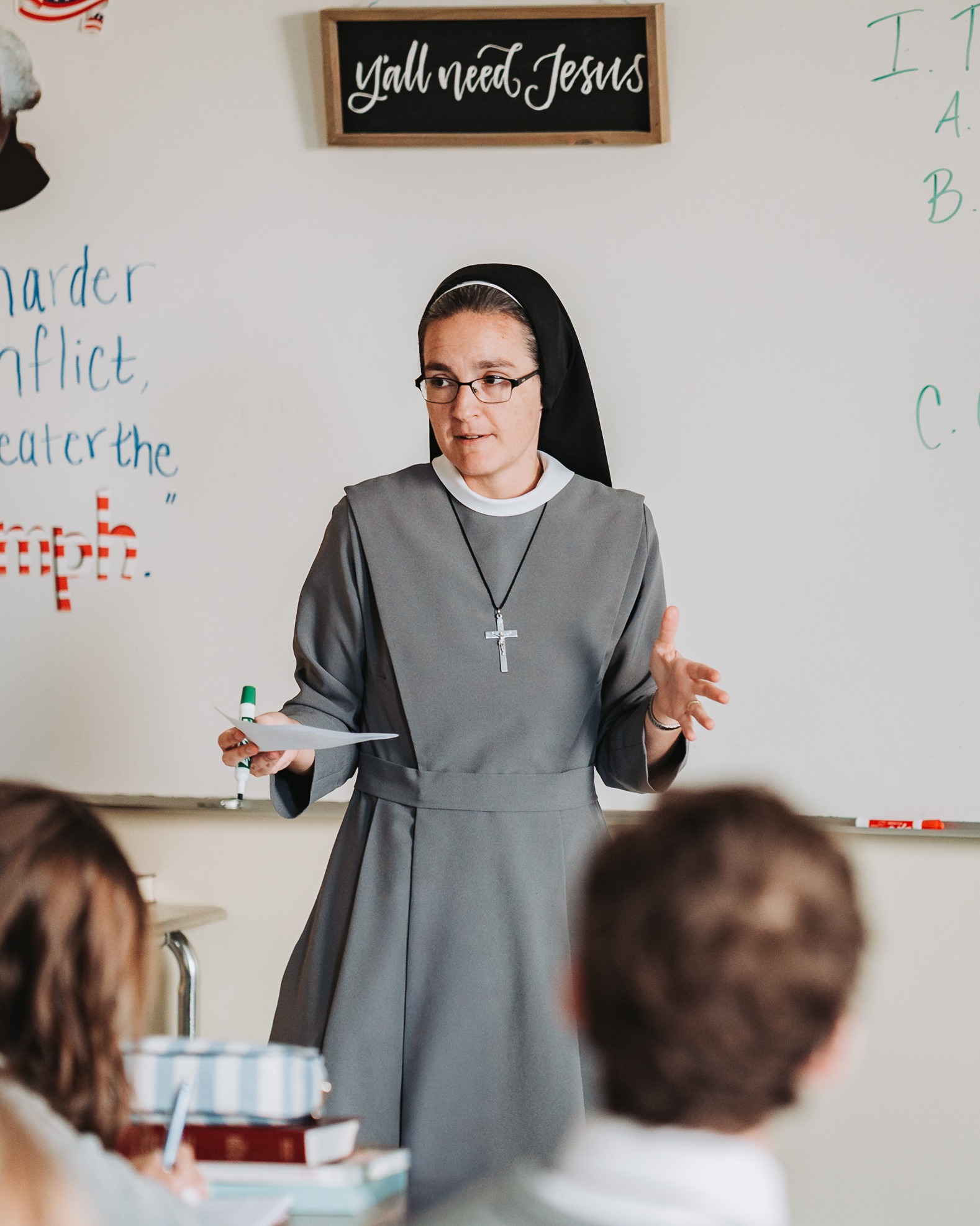 Nun in a gray habit and glasses teaching in a classroom with a whiteboard behind her.