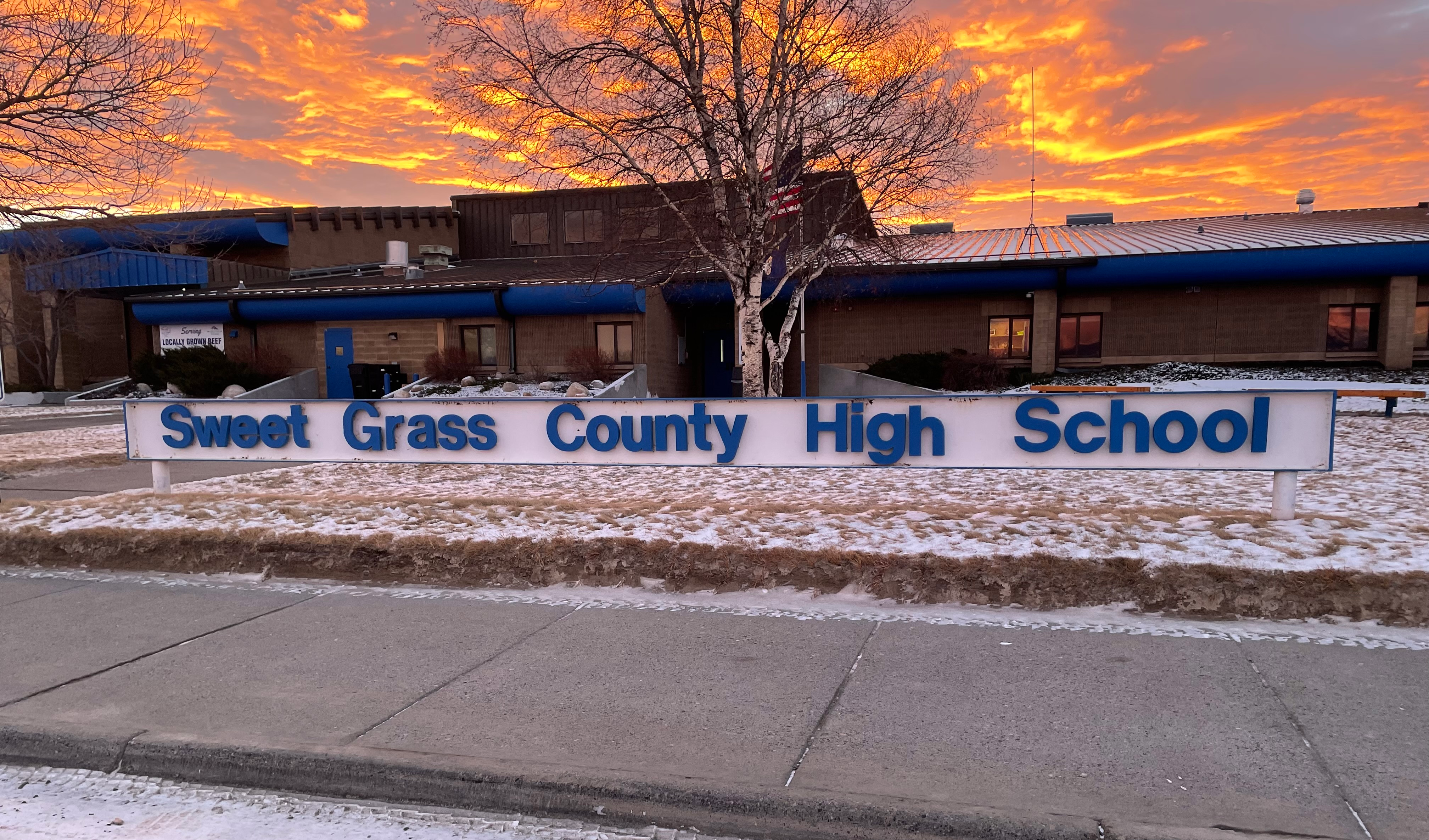 photo of the Sweet Grass County High School sign with the school building behind it and a colorful morning sunrise behind the building